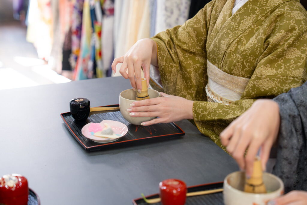 Hands whisking matcha at a table during a Kyoto tea ceremony experience, with wagashi sweets and tea utensils.
