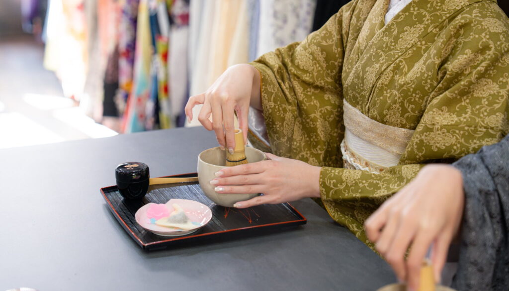 Hands whisking matcha at a table during a Kyoto tea ceremony experience, with wagashi sweets and tea utensils.