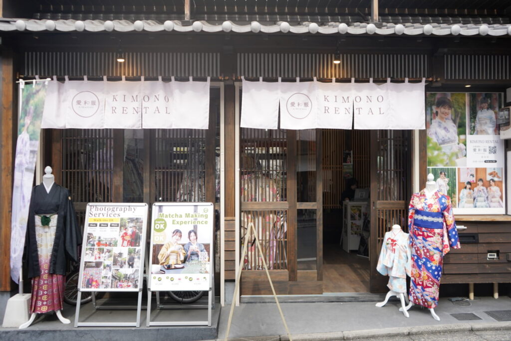 Exterior of a Kyoto kimono rental shop with “KIMONO RENTAL” noren curtains, mannequins in kimono, and signboards for photography and matcha making experiences.