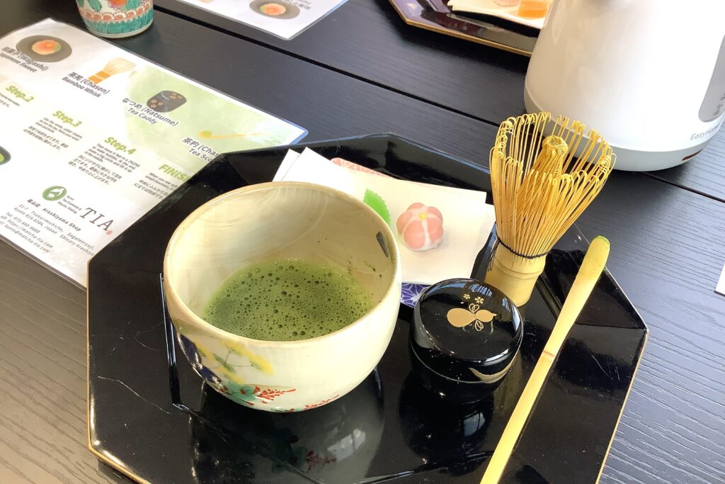Matcha tea set on a table with a ceramic bowl of freshly whisked matcha, bamboo whisk, tea caddy, and wagashi sweets at TIA Kyoto tea ceremony experience.